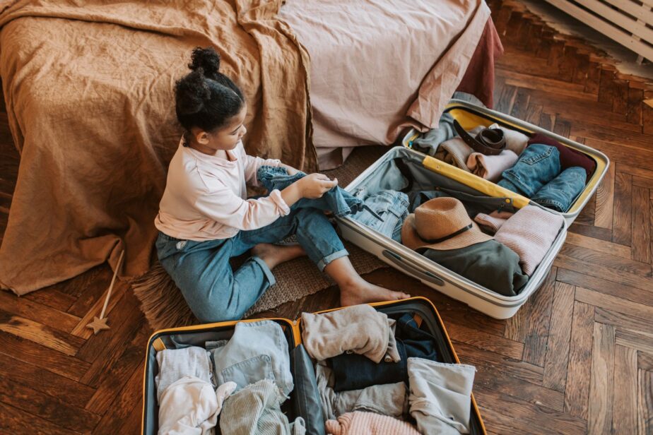 Une enfant prépare des vêtements dans une valise ouverte, assise sur un lit.