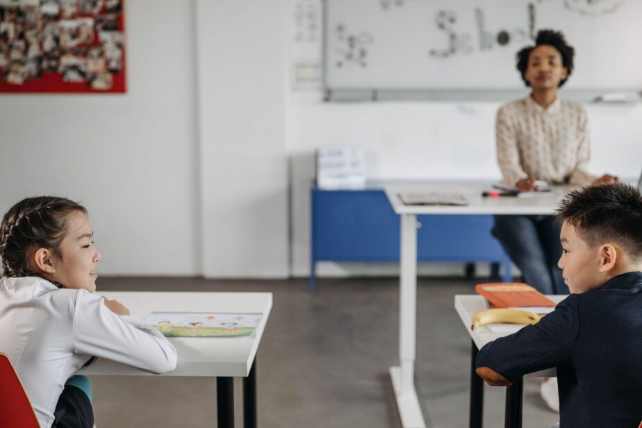 Deux élèves sont assis à leur table, avec une enseignante debout au fond de la salle de classe.